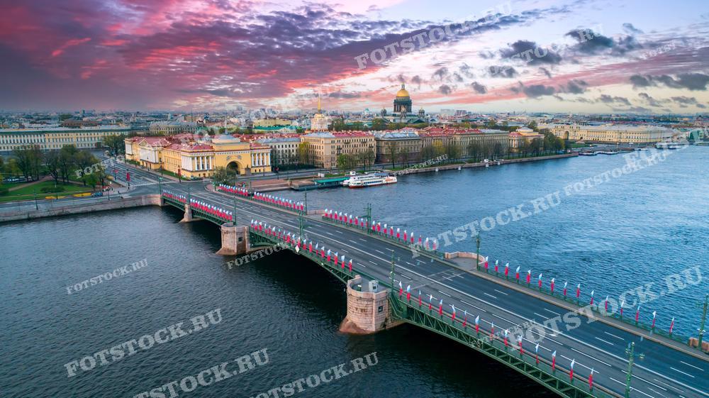 Эта фотография показывает вечерний или утренний вид Санкт-Петербурга. На фоне видны раскинувшийся город и изменчивое небо, окрашенное в теплые оттенки заката или рассвета. Изображение отлично подойдет для иллюстраций в путеводителях, исторических рассказах о городе, а также в качестве декоративного элемента для интерьеров, подчеркивающего красоту архитектуры и водных пространств Петербурга.